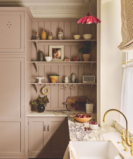 dusty pink lilac traditional shaker kitchen with marble countertops, open shelving with vintage decor, a cafe curtain window and roman blind and a sink with brass faucet