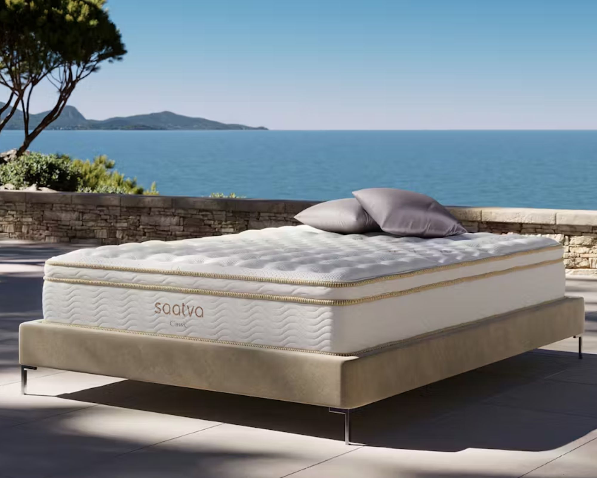 A Saatva Classic Mattress pictured outdoors with two grey cushions on top, and a bright and sunny seascape behind.