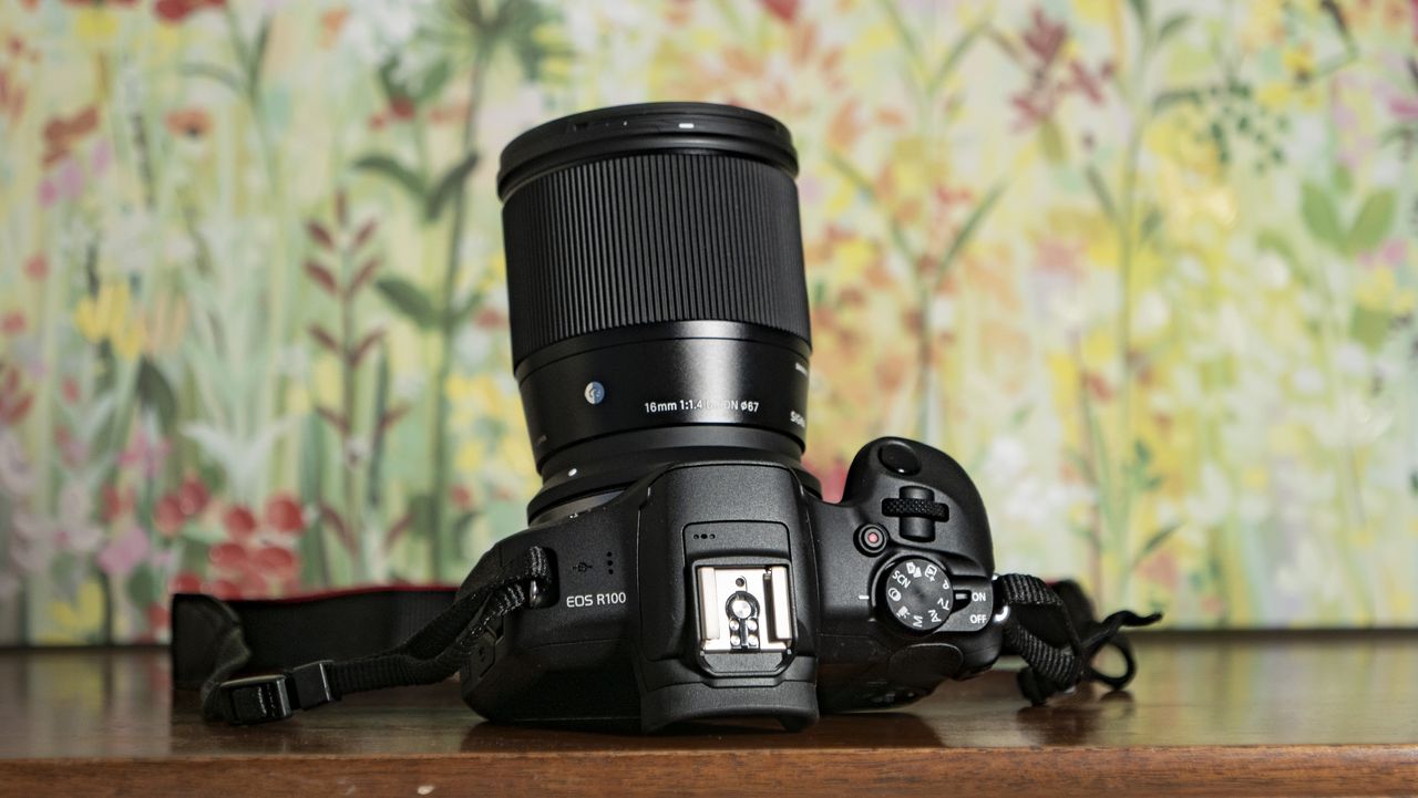Sigma 16mm F1.4 DC DN Contemporary lens attached to a Canon EOS R100, on a wooden surface with floral backrop