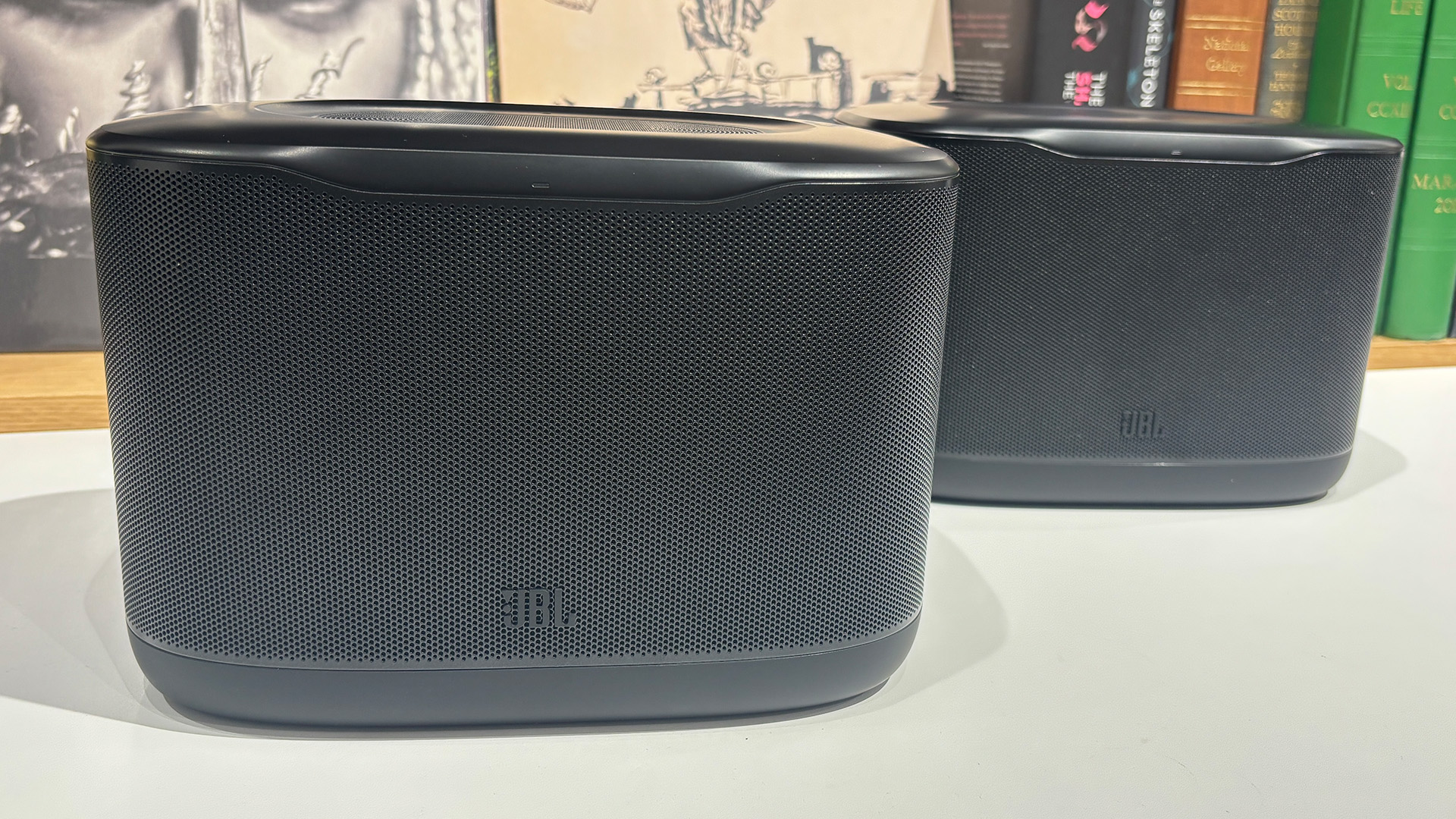JBL Cove X1 multi-room wireless speakers on white table in front of busy bookshelf