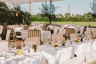 The outdoor space of a chic French restaurant features mid-century modern decor including beige soft seating, all overlooking a green Parisian park and with the Eiffel Tour in the distance.