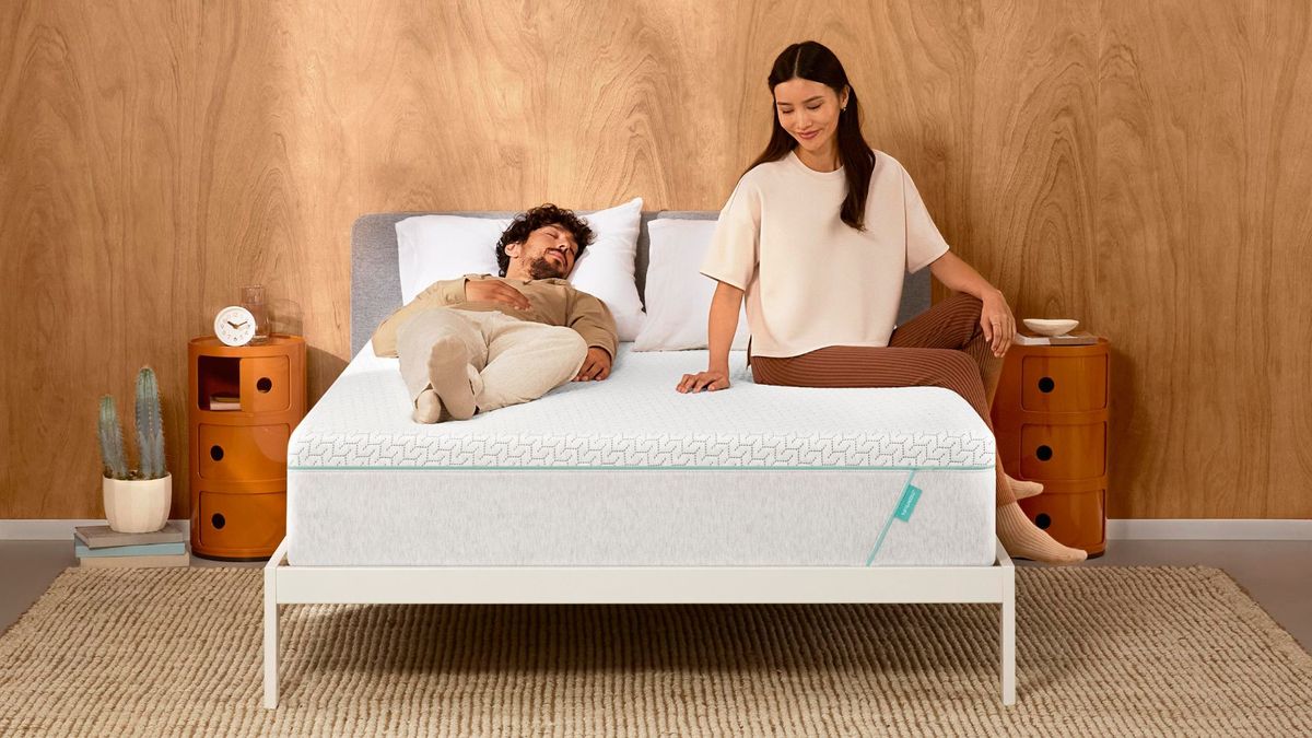 A couple sit on top of the Tuft &amp; Needle Original Hybrid mattress, which is placed on a white bed frame in a neutrally decorated bedroom