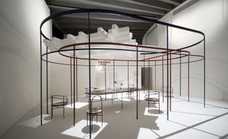 View of Librizzi’s room installation - a red and blue oval shaped frame plus a table, chairs and a large, white hashtag style structure above. The walls and flooring around the frame are light grey and black text can be seen on one wall