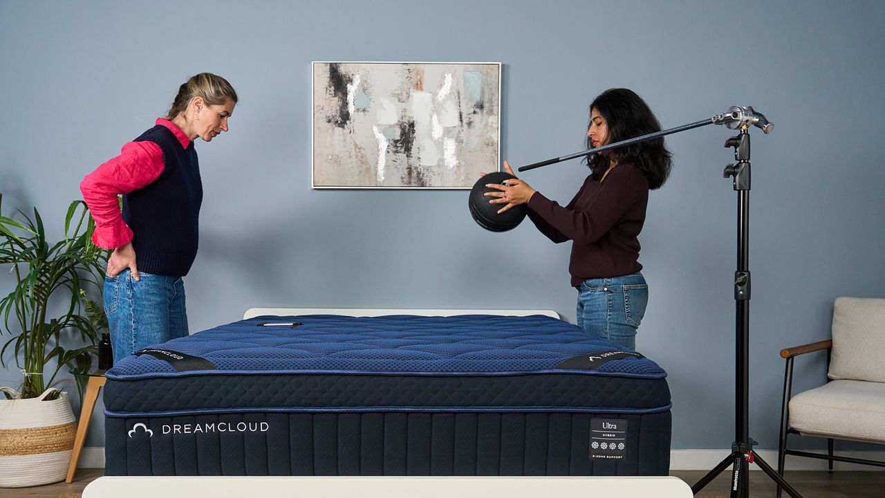 Two mattress testers stand above a blue mattress, with one dropping a 5kg slam ball onto its surface while another records its reading on her phone. This helps measure motion isolation.