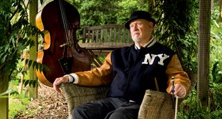 Danny Thompson wears a NY varsity jacket and pork pie hat, and in a garden chair, holding his bow. His upright bass is in the background.
