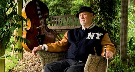 Danny Thompson wears a NY varsity jacket and pork pie hat, and in a garden chair, holding his bow. His upright bass is in the background.
