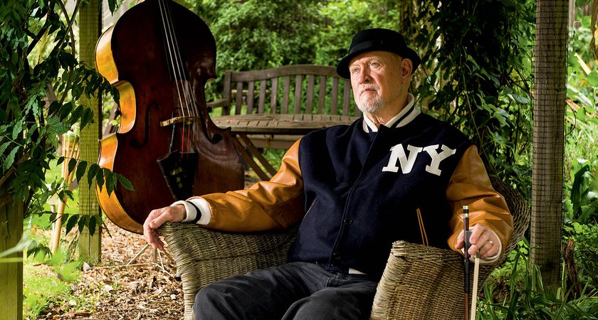 Danny Thompson wears a NY varsity jacket and pork pie hat, and in a garden chair, holding his bow. His upright bass is in the background.