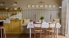 A vintage style kitchen with yellow cabinets, vintage tiling, a dining table with white tablecloth and wooden dining chairs, decorative plates and wood panelling.