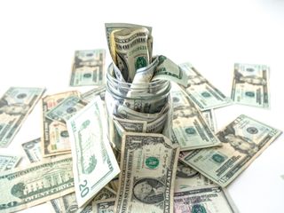 A closeup photograph of United States cash or paper bills overflowing out of a glass mason savings jar and onto a white background.