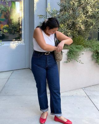 influencer Marina Torres poses in a warm weather outfit with a claw hair clip, white tank top, basket bag, belted high waist cuffed jeans, and red ballet flats