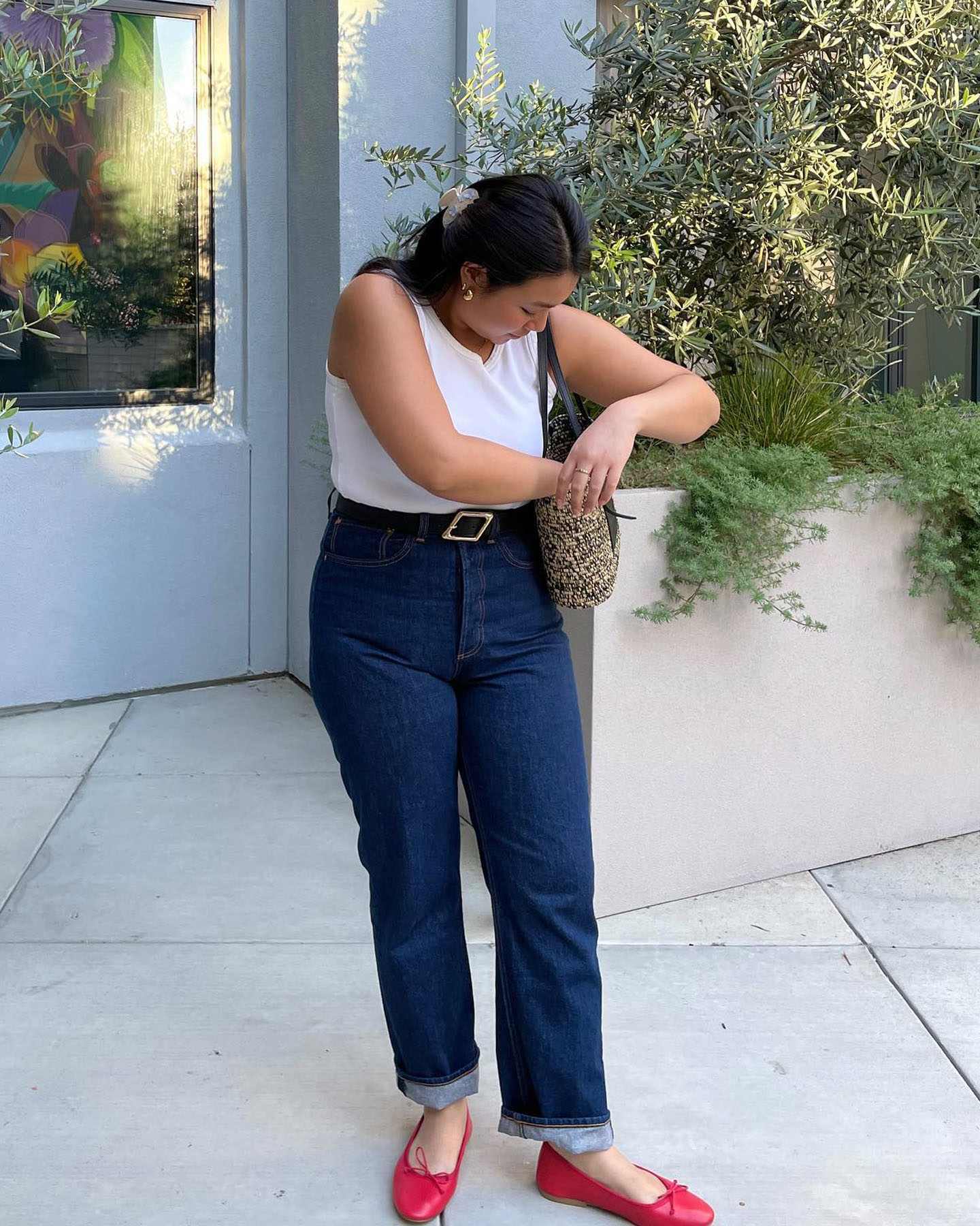influencer Marina Torres poses in a warm-weather outfit with a claw hair clip, white tank top, basket bag, belted high-waist cuffed jeans, and red ballet flats