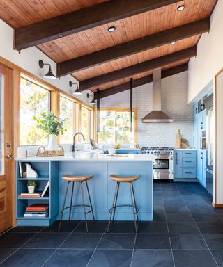 Bright kitchen with bright blue cabinetry, wooden ceiling, bar stool seating, gray tiled flooring, wall lamps, large floor to ceiling windows, white tiles on walls