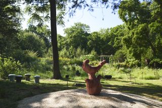 A concrete freestanding fountain with organic shapes painted in red, created by Rogan Gregory and photographed among greenery in a garden in East Hampton