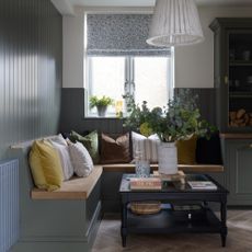 A corner with built-in wooden banquette seating painted sage green to match the wall panelling and a window above dressed with a floral-print Roman blind