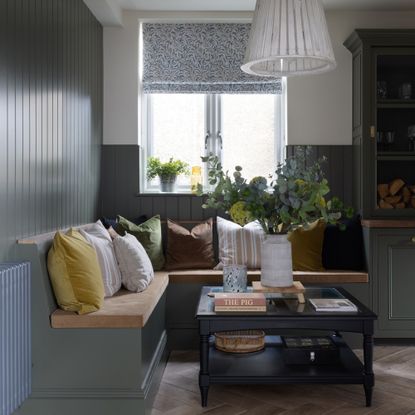 A corner with built-in wooden banquette seating painted sage green to match the wall panelling and a window above dressed with a floral-print Roman blind