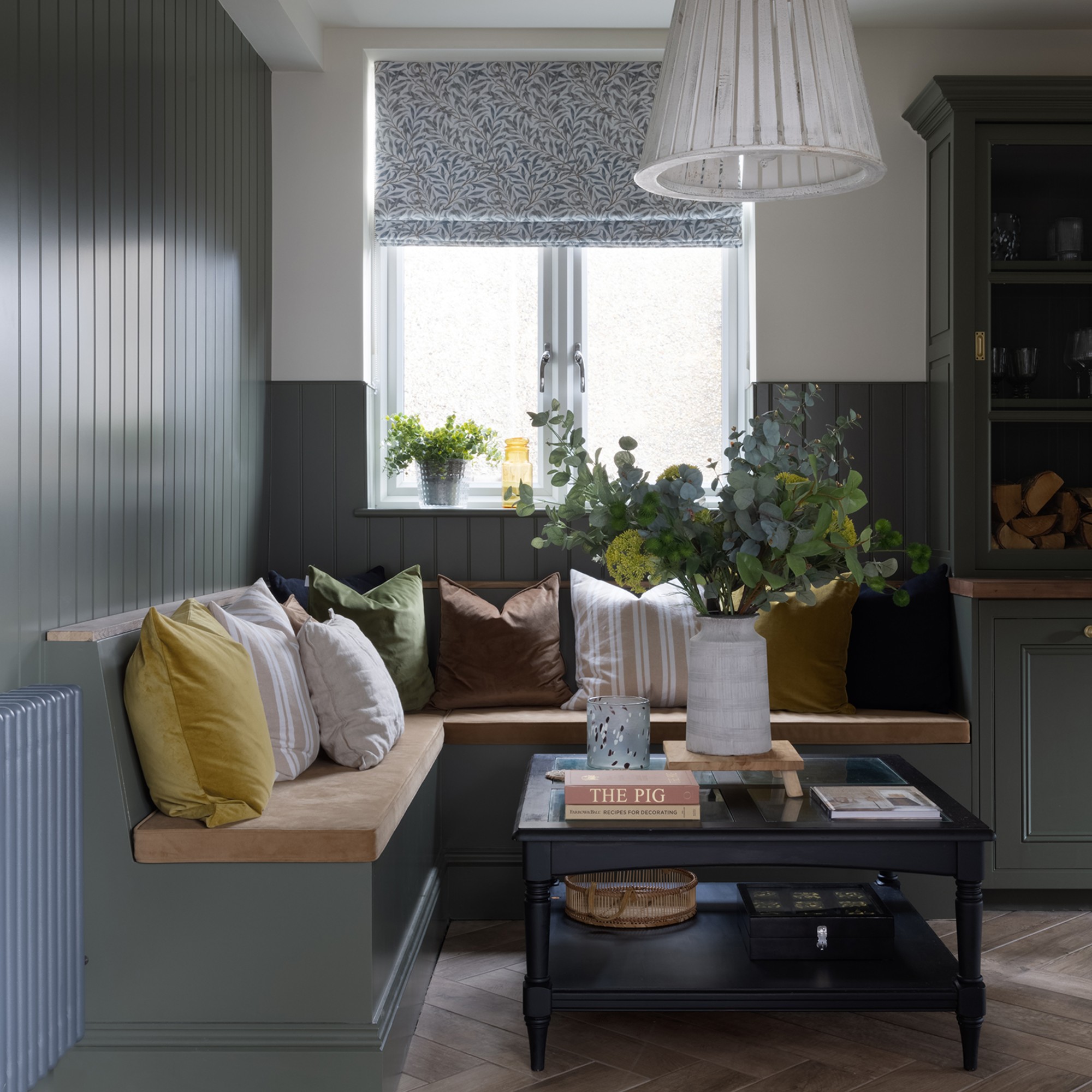 A corner with built-in wooden banquette seating painted sage green to match the wall panelling and a window above dressed with a floral-print Roman blind