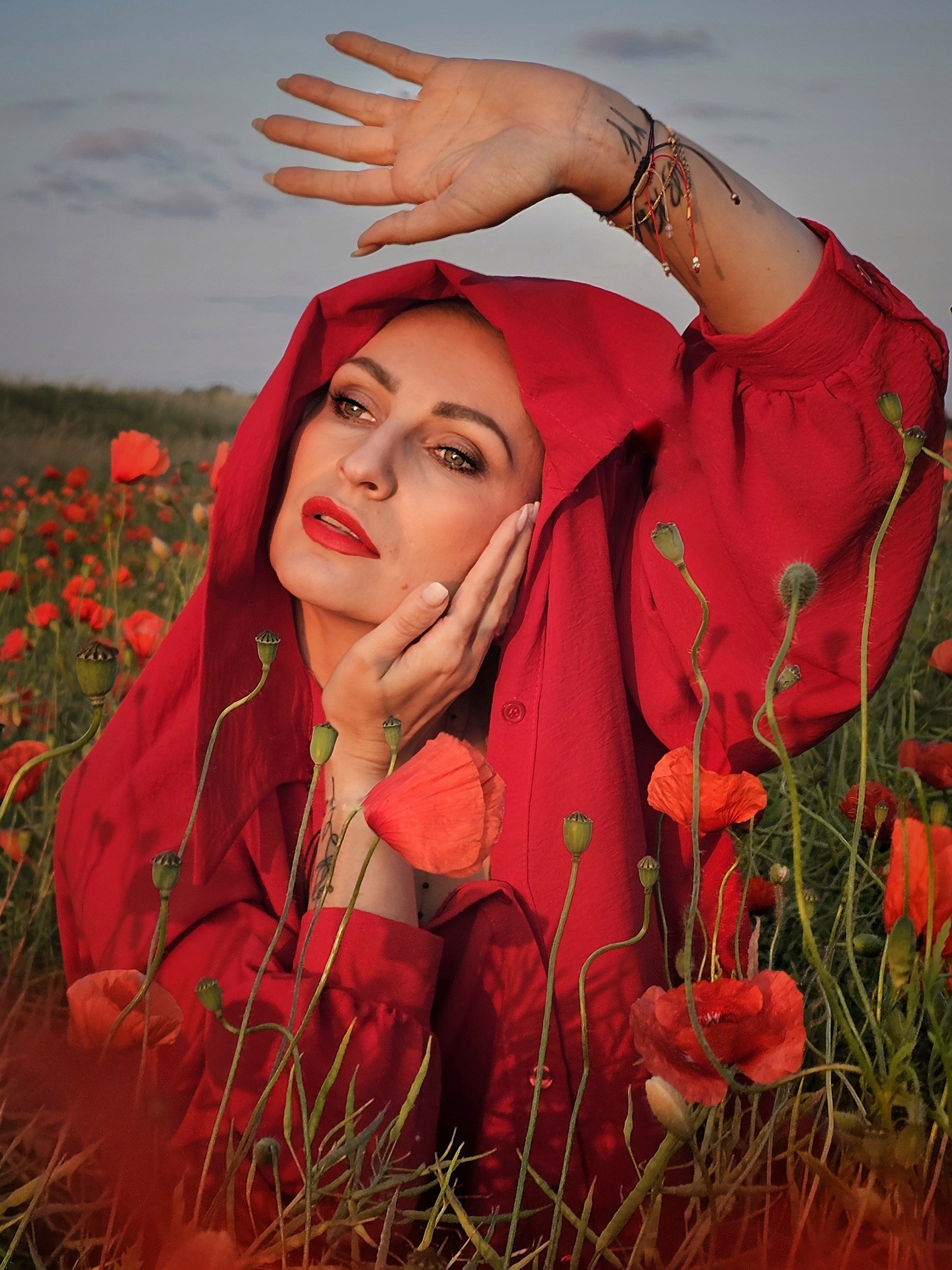 Woman in red cloak poses in a field of vibrant poppies at sunset, hand raised, amidst the blossoms