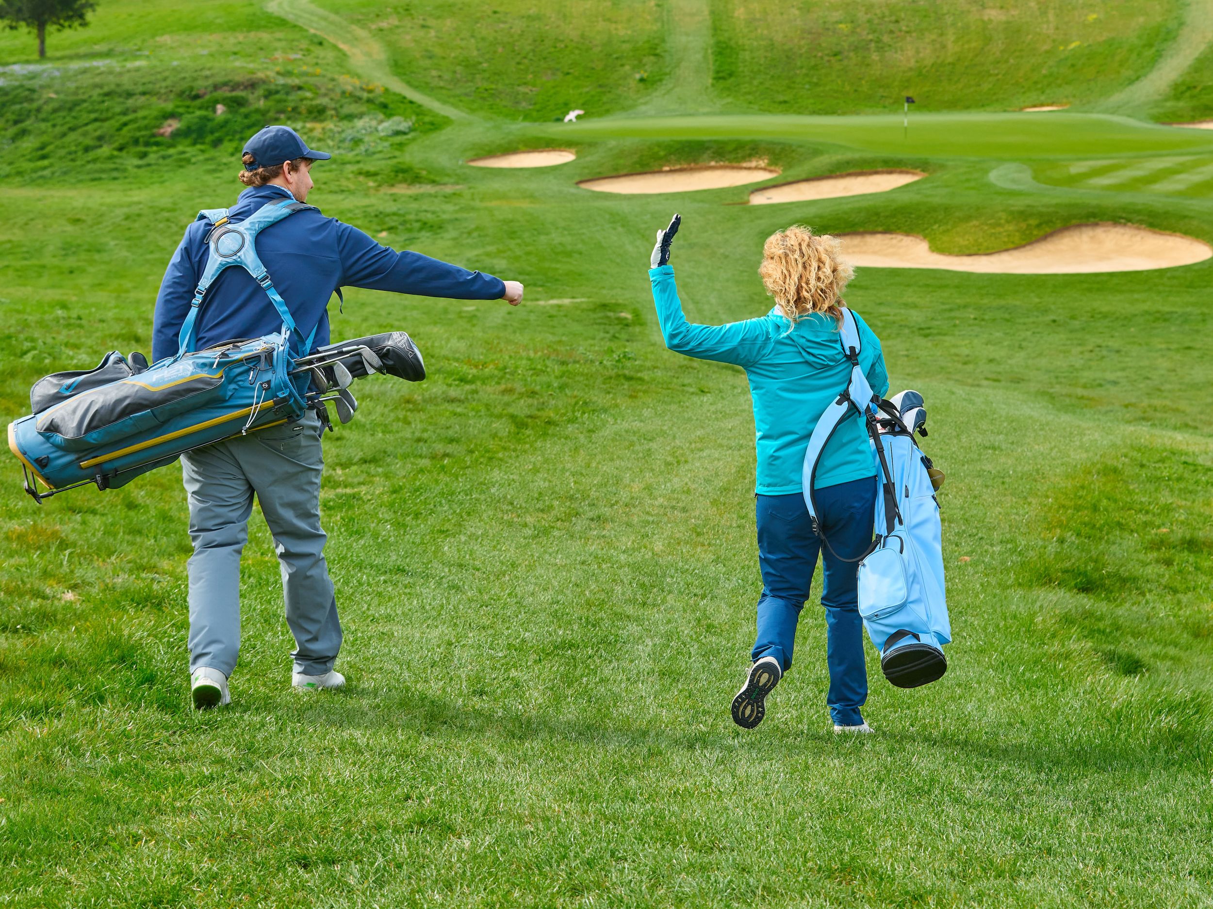 Two golfers walking towards a fairway. One has made a first and is offering it for a fist pump but the other golfer is waving this invitation away
