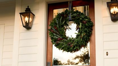 A front door Christmas wreath hanging by a ribbon from the top of a wooden door with a glass window. 