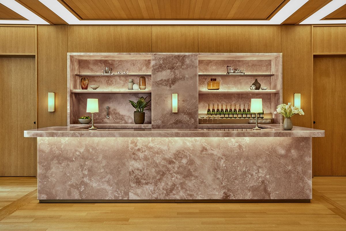A hotel salon bar features perfectly symmetric, interiors including a striking, rose-shaded marble counter and shelving unit, yellow-hued table lamps, and paneled wooden walls.