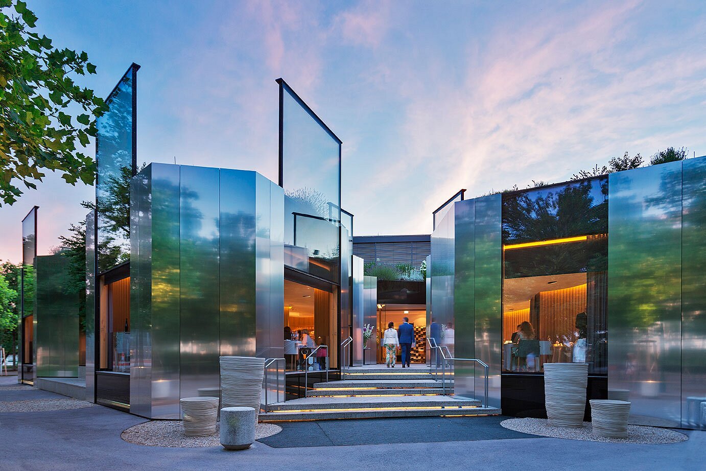 The mirror and glass, multi-part futuristic structure of a restaurant sited in a park is brought to life by a pink-toned sunset, clouds in the sky, and plants, as diners enjoy their meal absorbed in a cozy, orange atmosphere.