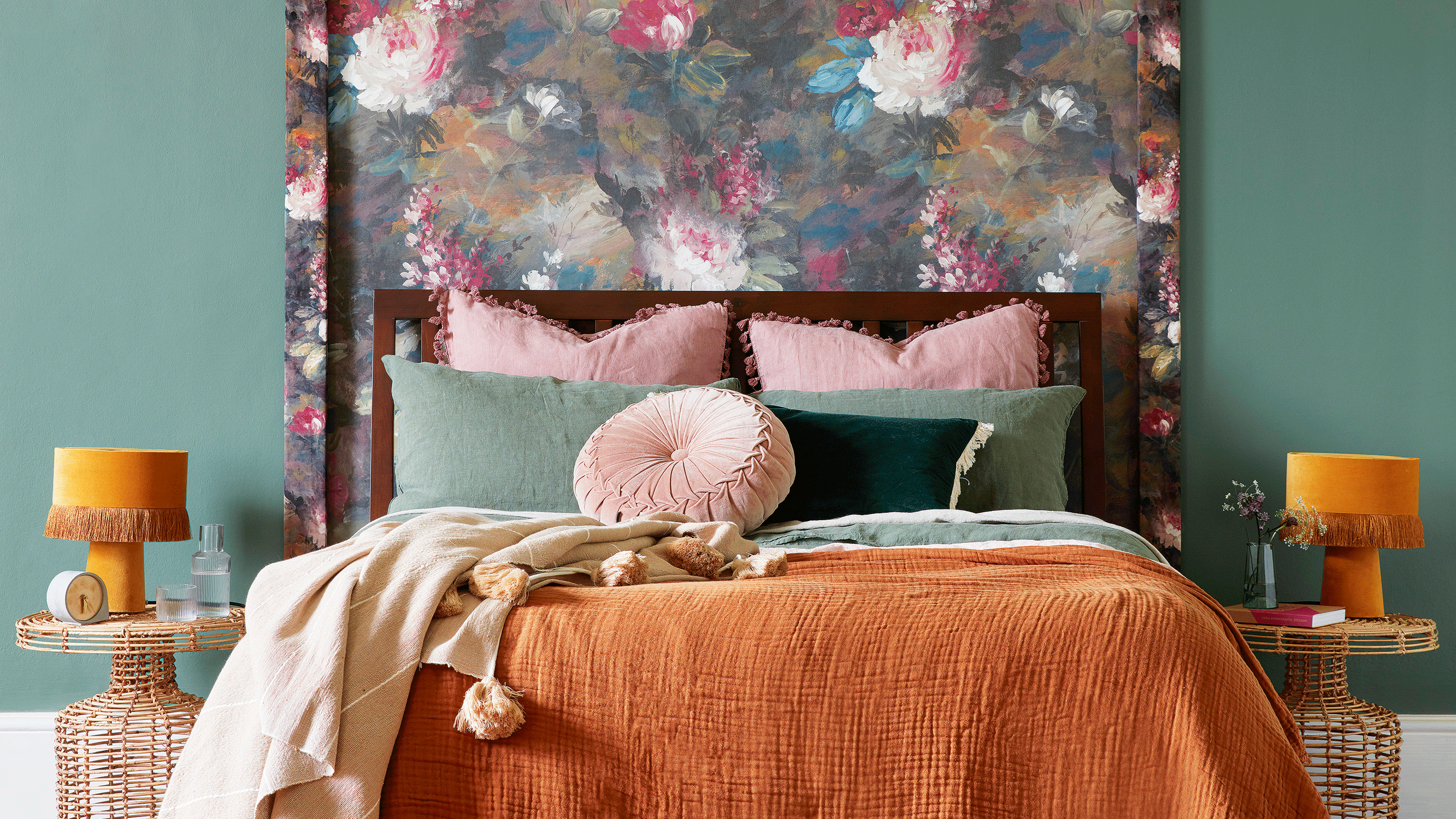 Teal bedroom with floral wallpaper behind bed and orange bedding and lamps