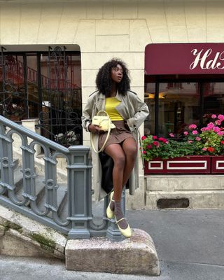 Emmanulle wears a tan trench coat, yellow top, chocolate brown skirt, ivory bag, brown socks, and ivory mary janes.