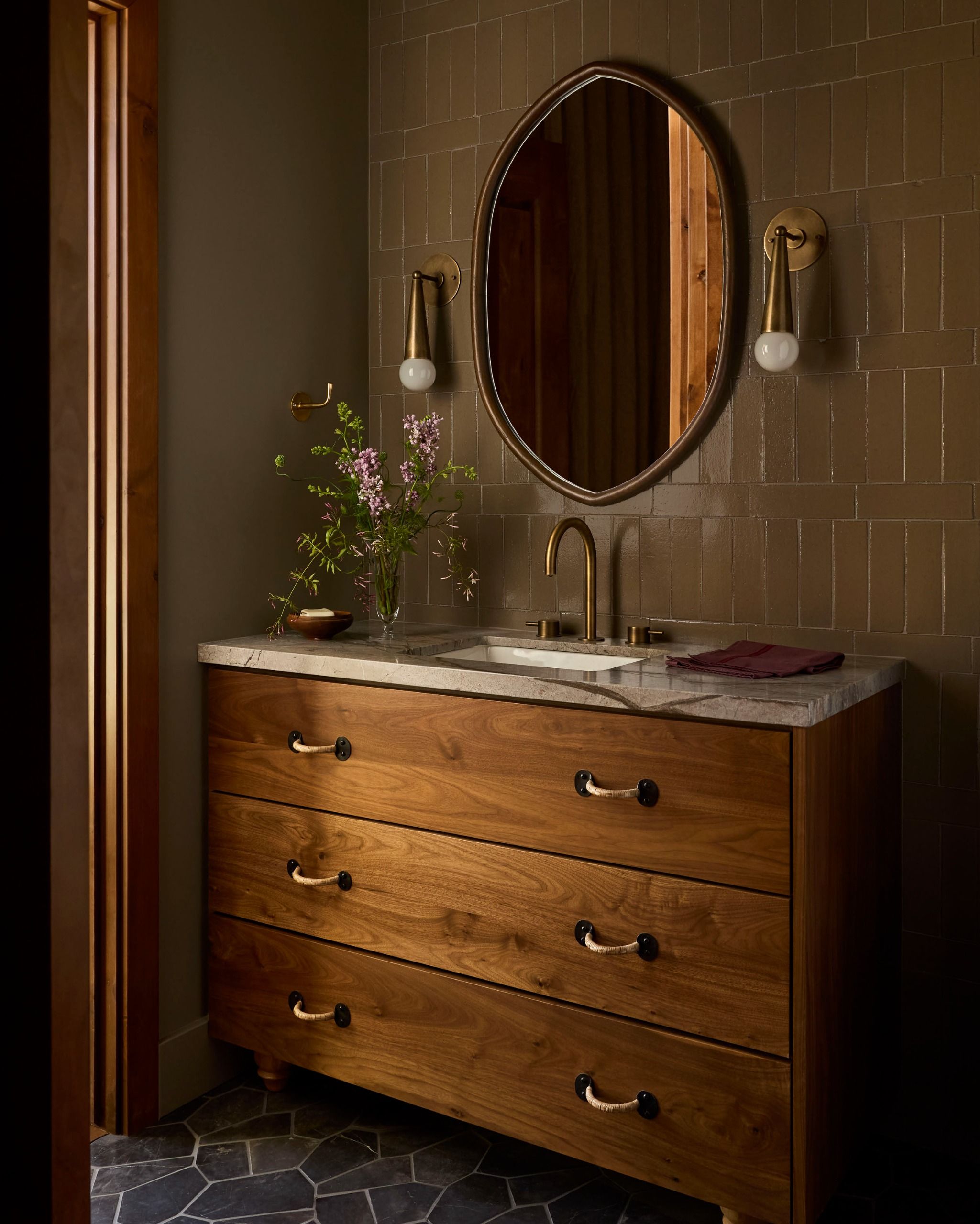 A bathroom with a vanity unit with three wooden drawers with brass handles and an oval mirror above it with a wall light either side and a vase of flowers by the sink