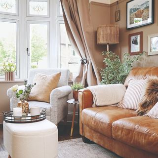 A small living room painted in a light brown with a gallery wall, a brown leather sofa with contrasting cushions and a cream rug underneath