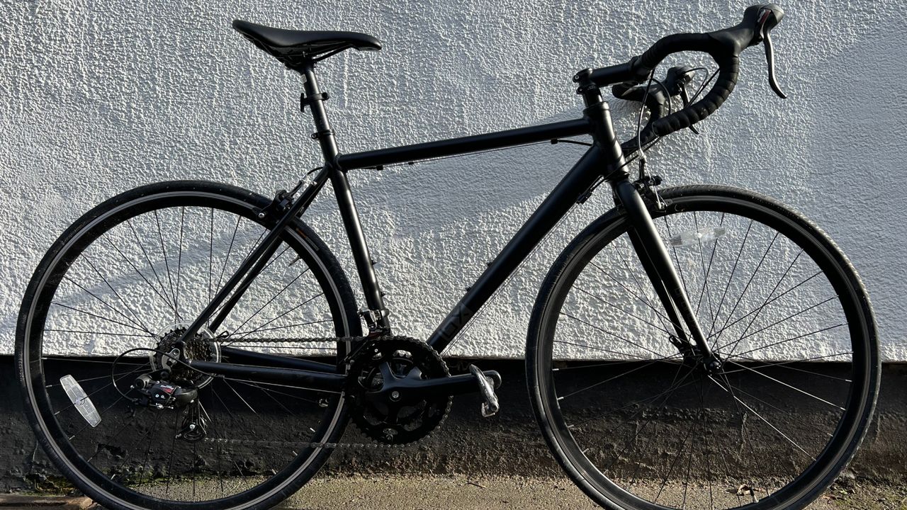 The black Brand X Road bike in full leaning against a mostly white wall 