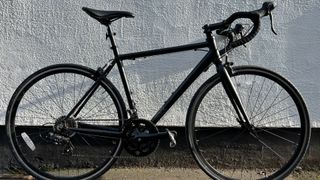 The black Brand X Road bike in full leaning against a mostly white wall