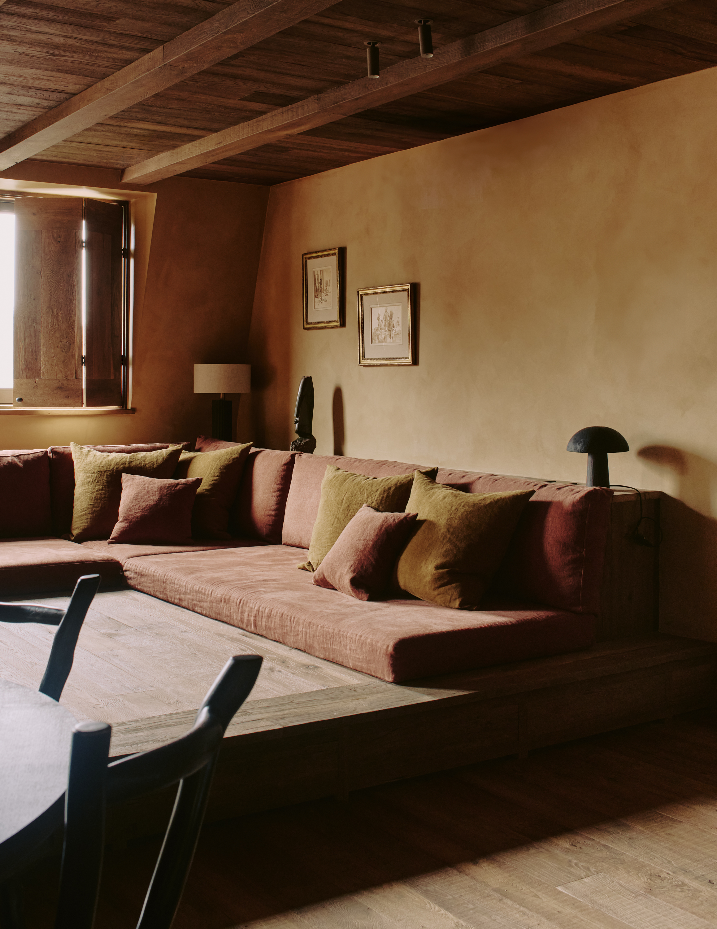 Image of an orange limewash living room with a sectional sofa that's on a raised part of the floor. There are wooden ceilings and a window with wooden shutters.