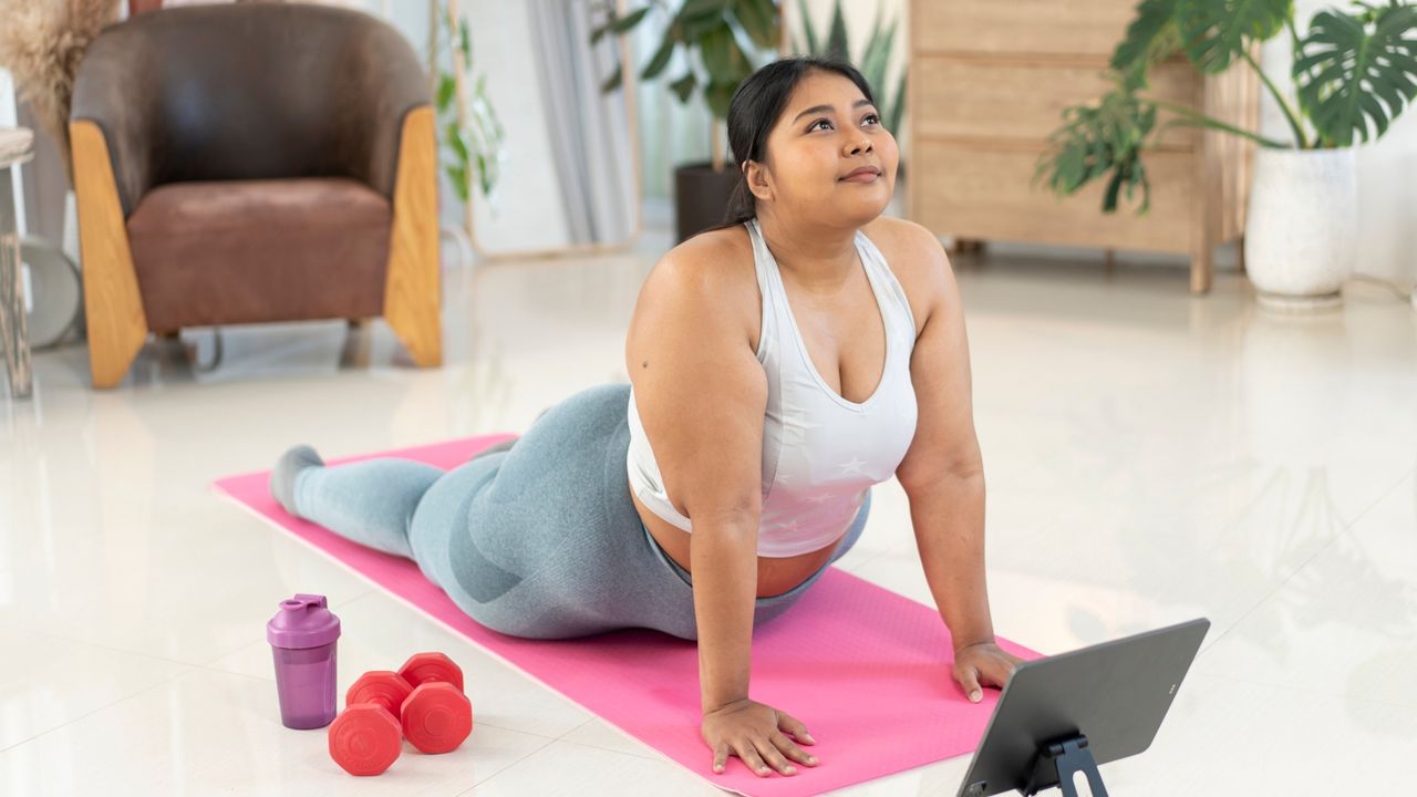 woman wearing white crop top and light blue leggings doing a spinal extension pilates pose on a pink mat facing the camera in a living room setting. 