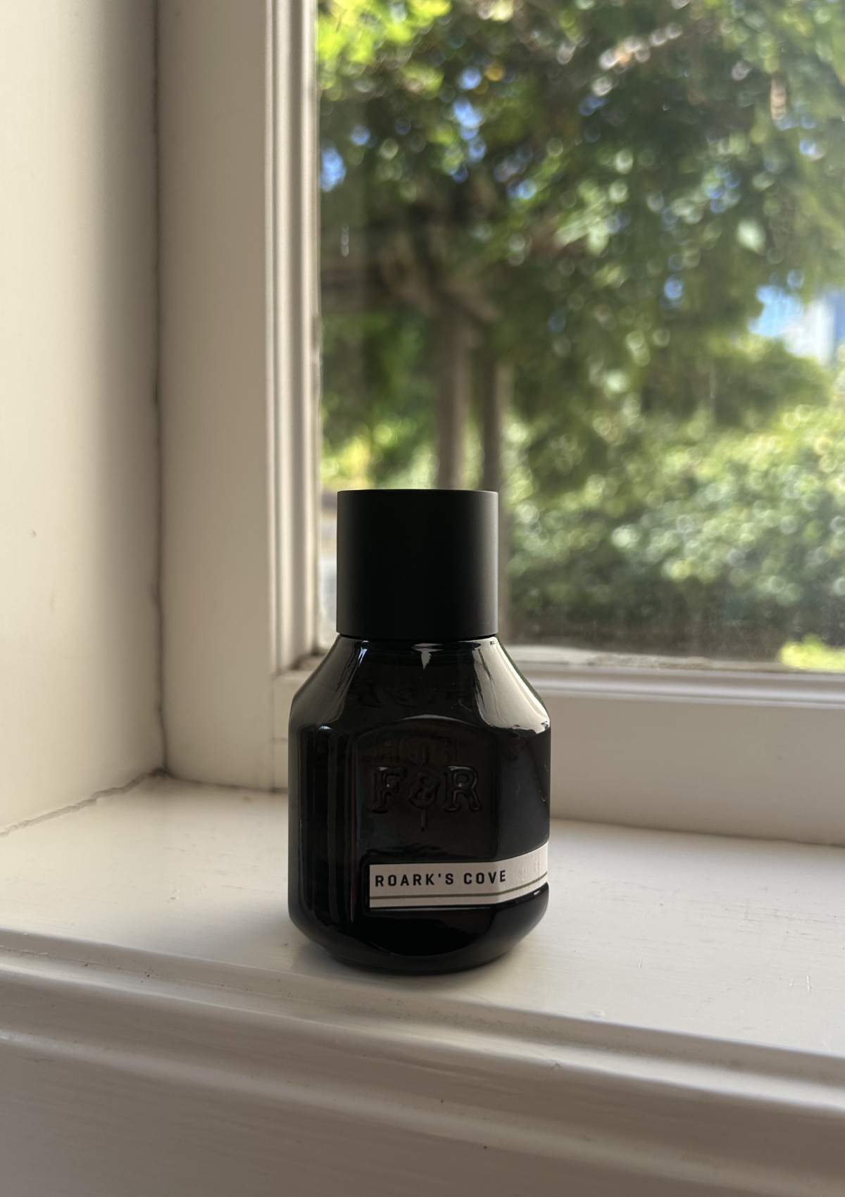 Roark&amp;rsquo;s Cove perfume by F&amp;amp;R in a dark glass bottle on the windowsill, bright green trees blurred outside