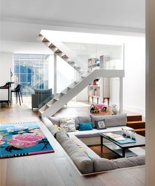 All white basement living room, gray sunken sofa, central staircase, dark wood flooring with pink and blue rug, black coffee table, glass panels, cabinets and shelves with books, black bar stools