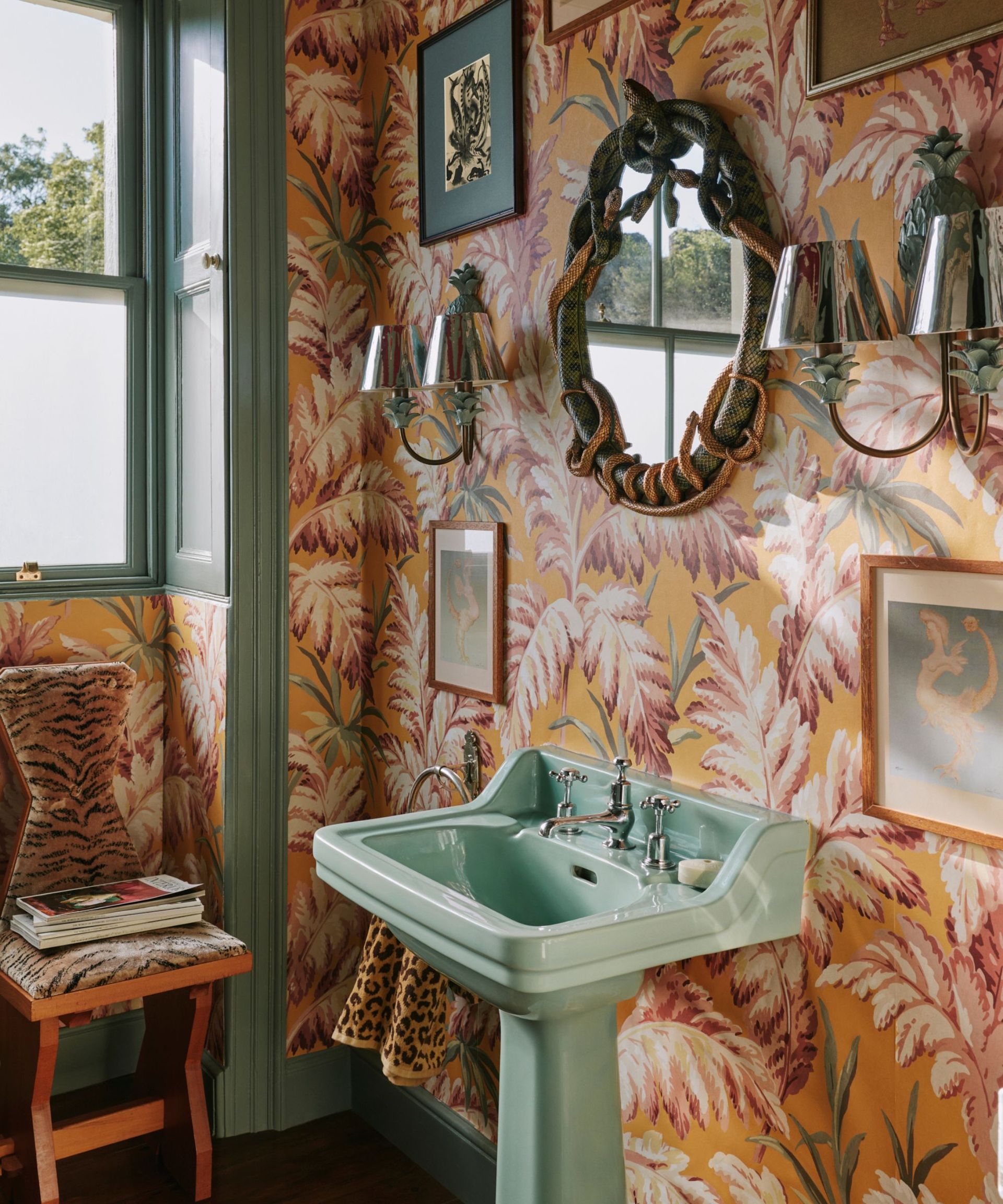 A small bathroom with and orange and pink botanical wallpaper, mint green pedestal sink, and vintage wall decor
