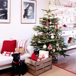 A living room with a white rattan-base sofa and a decorated Christmas tree with a wooden crate next to it filled with presents