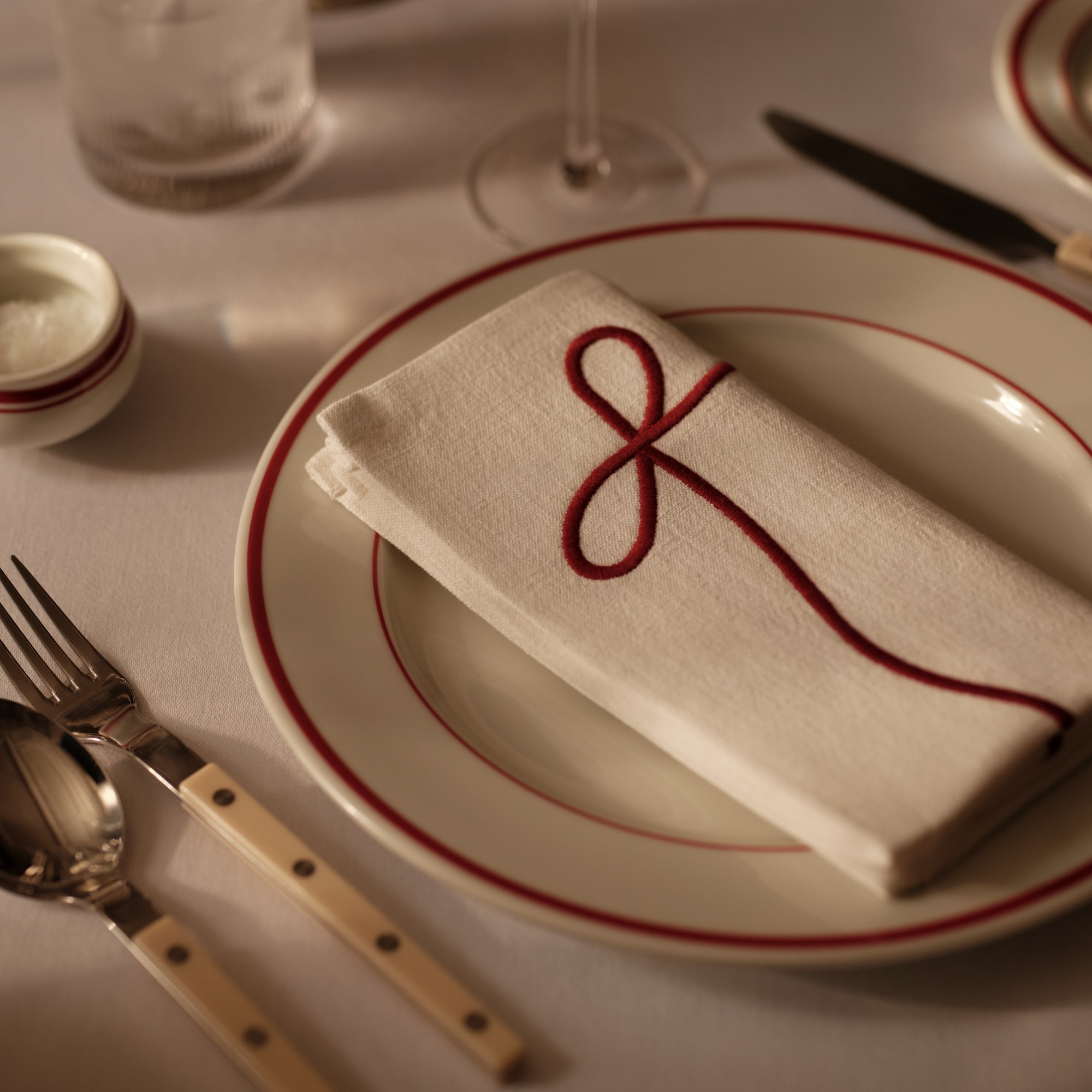 A set table with a bistro plate in white with a red trim and a red bow-embroidered white napkin on top of it