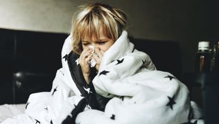 a young child with blonde hair sits on a bed covered in a blanket while blowing his nose into a tissue.