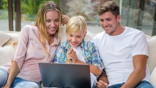 A woman, a child, and a man looking happily at a laptop display while seated on a couch.