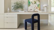 A white home office with a deep blue chair, a white desk, a vase of stems, a leaning print, a white mushroom lamp, and a stack of drawers