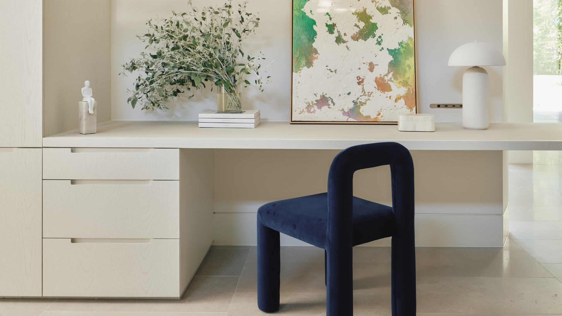 A white home office with a deep blue chair, a white desk, a vase of stems, a leaning print, a white mushroom lamp, and a stack of drawers