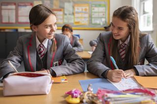 Izabella Cresci as young Birdy and Daisy Jacob as young Maggie, in their school uniforms in a classroom, doodling in their schoolbooks and smiling at each other
