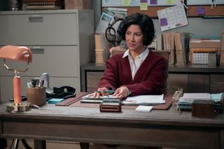 Stephanie Beatriz as Didi, sitting at her office desk and holding a pen, in episode 107 of A Man on the Inside.