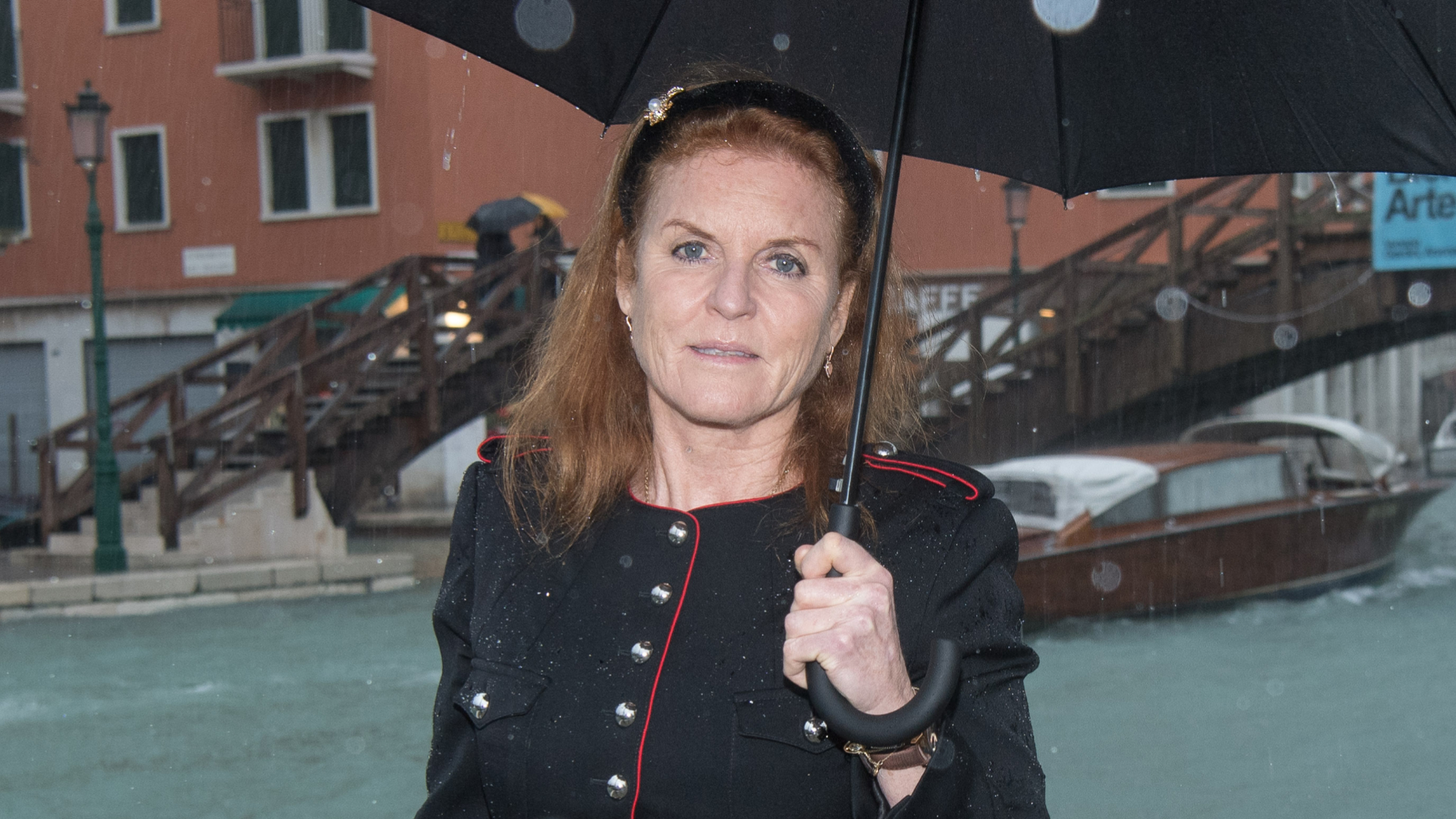 Sarah Ferguson wears a black jacket with metal buttons and red piping and holds a black umbrella while standing in the rain in Venice