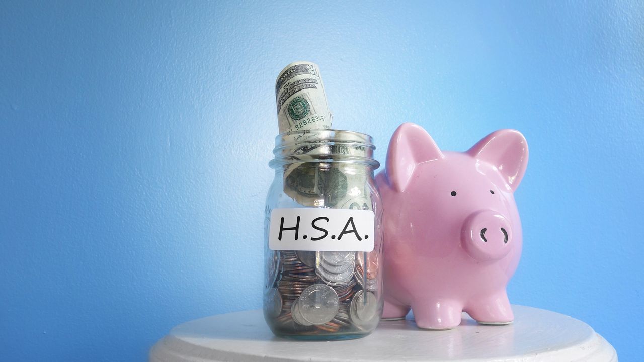 A piggy bank sits next to a jar labeled HSA that&#039;s filled with cash.