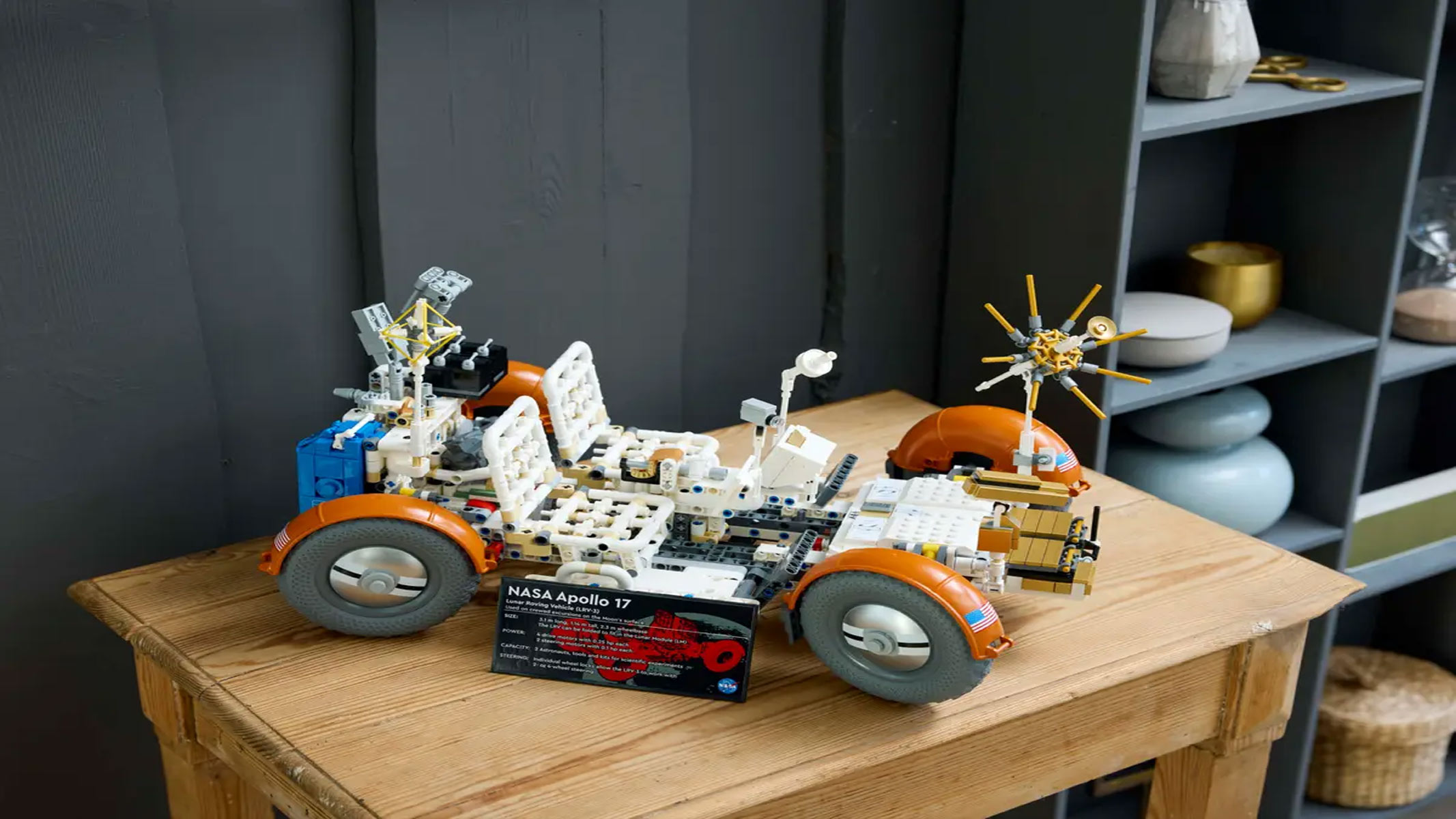 A brick-built vehicle with four wheels is placed behind a display plaque with some information on, all on a light brown wooden surface.