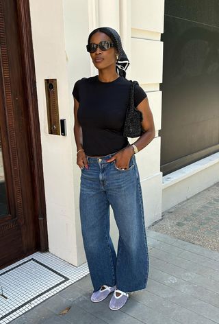 A woman standing outside wearing a black t-shirt with barrel-leg denim and white mesh flats.