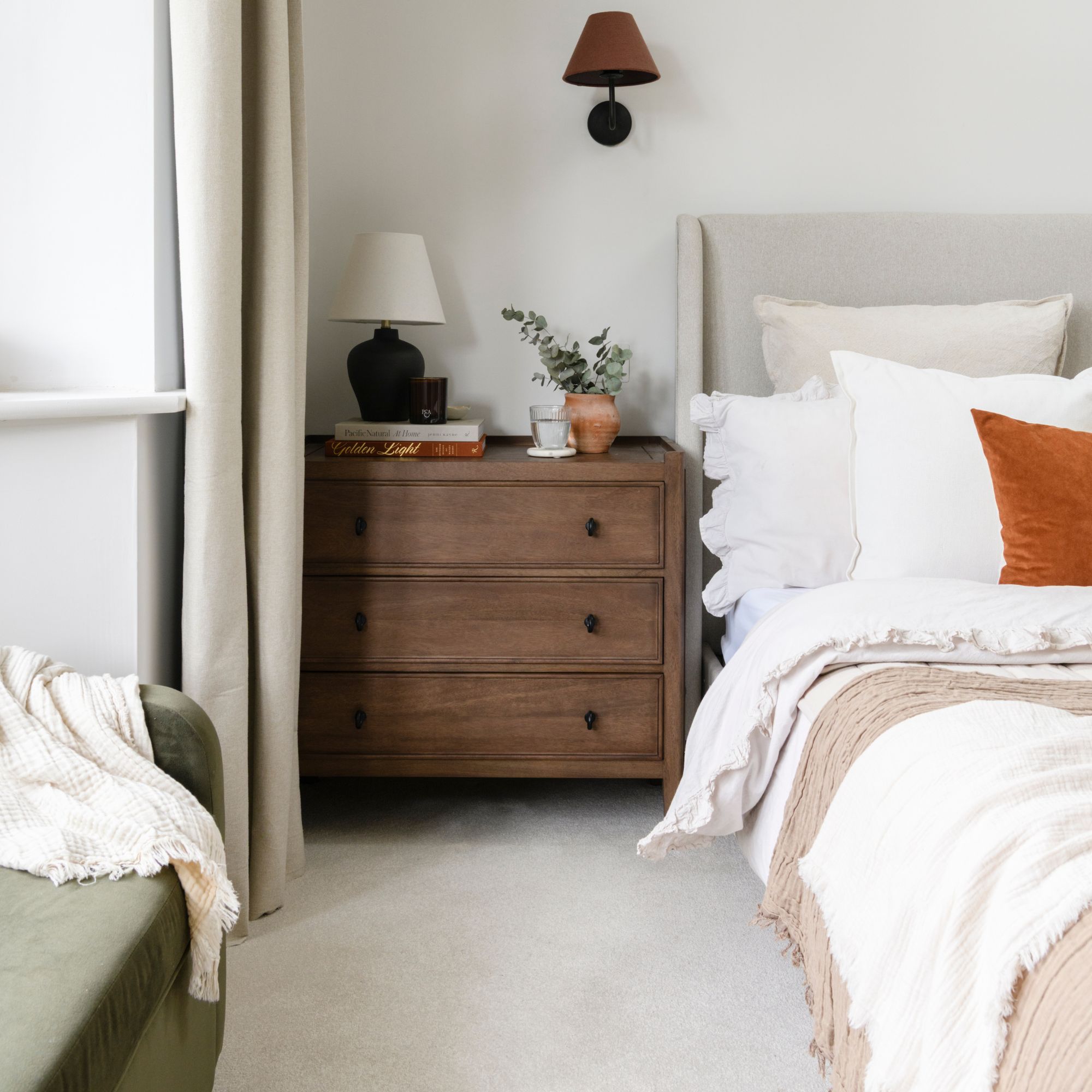 White painted bedroom with beige carpets, white bedding on the bed, and a wooden chest of drawers next to it