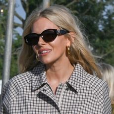 Sienna Miller wearing sunglasses, gold earrings, a plaid sporty jacket, and white linen pants, with wellies at Glastonbury. 